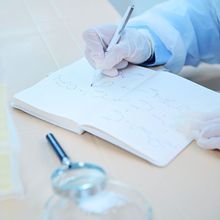 A scientist with gloved hands sitting at a table with a magnifying glass and lab glassware and writing in a notepad with a pen.