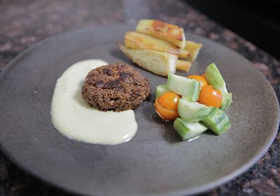 Photo of a brown soy pulp patty sitting in a cream sauce alongside a cucumber and tomato salad and baked yams.