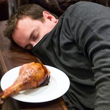 A photograph of a man sleeping on a couch next to a plate with a poultry leg on it.