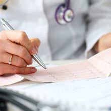 A white coat-wearing doctor holds the results of an EKG test in one hand and traces over them with a pen in the other hand