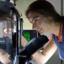 Alma Dal Co wears a blue shirt and red necklace and uses a pointer to examine a component of a laboratory machine.&nbsp;