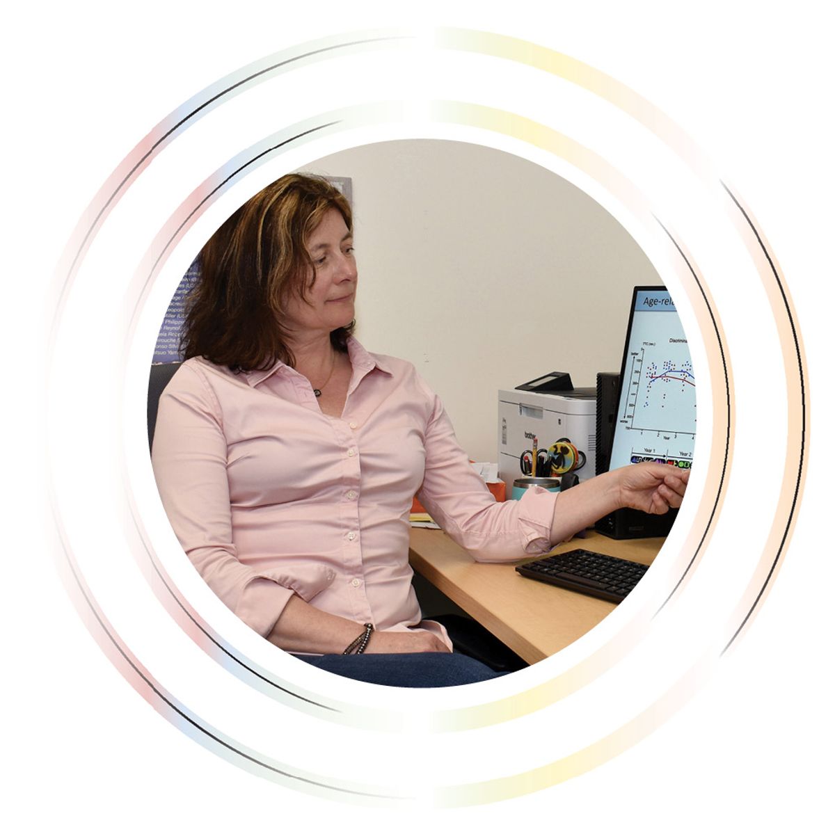 A woman sits at her desk while gesturing to an image on the computer screen next to her.