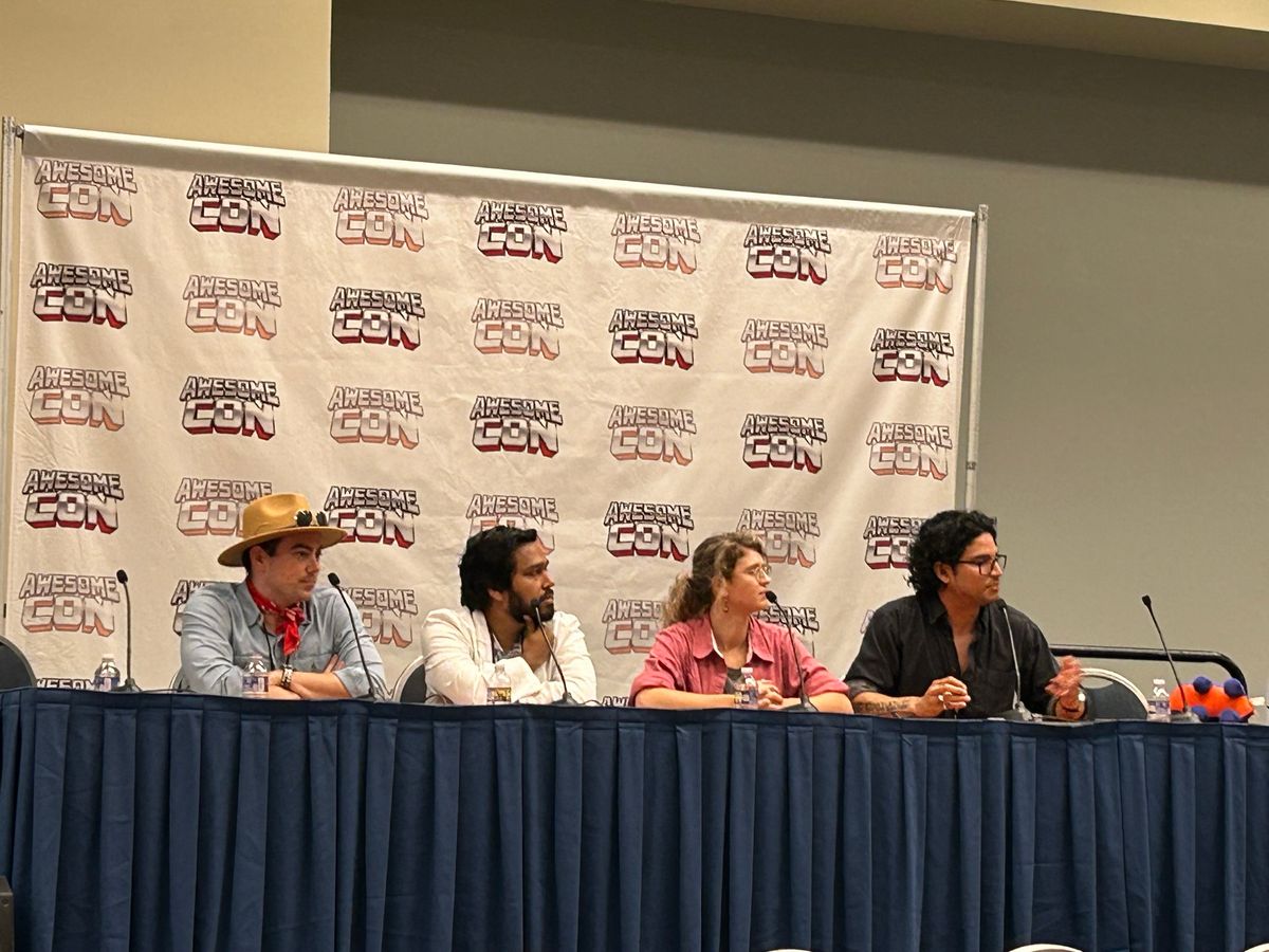 Carlo Quintanilla (far right) speaks at a panel table for a science-fiction conference, joined with three other panelists. He has dark hair and is wearing a dark gray shirt.