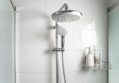 Image of a showerhead with running water. There is a soap dispenser and toothbrush on a wire shelf in the shower.