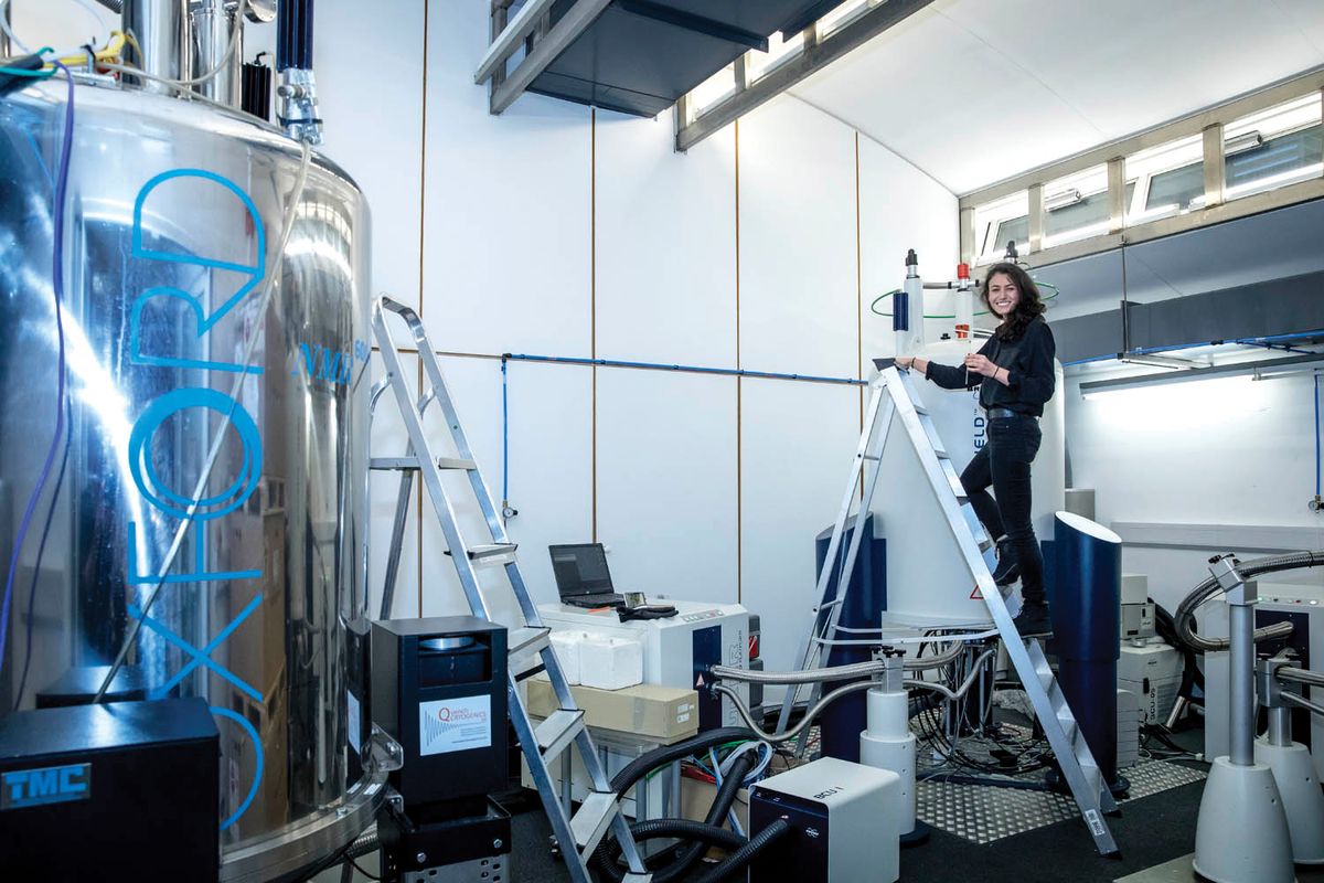 Photo of Gabriella Heller standing on a ladder next to a nuclear magnetic resonance spectrometer.