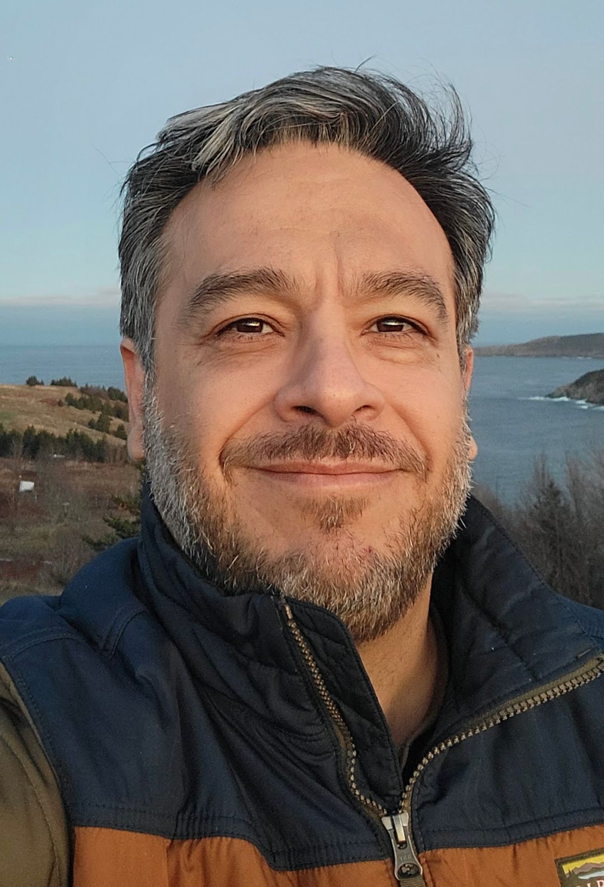 A headshot Mehrdad Hajibabaei smiling in front of a shoreline.