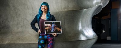 Beata Mierzwa, a postdoctoral researcher at University of California San Diego and the founder of Beata Science Art, poses for a photograph holding a print of one of her science illustrations. She has bright blue hair and is wearing leggings from her microscopy print design that features blue and green and red and blue cells.