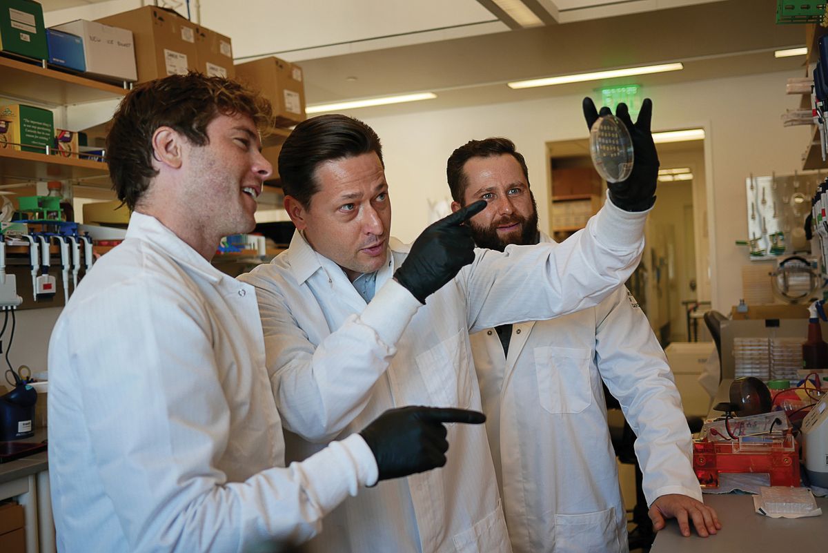 Three men are in a laboratory. They wear laboratory coats and are looking at a culture dish.