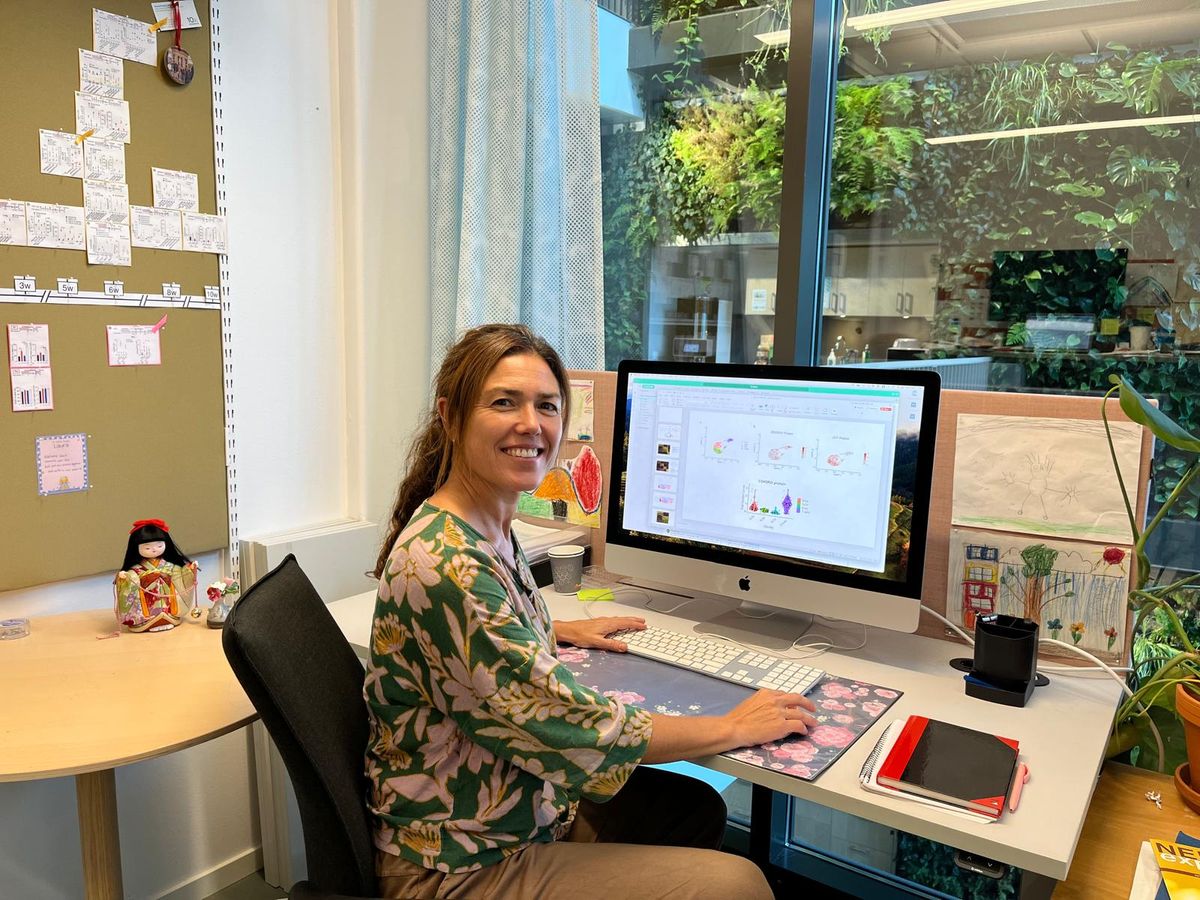 Itziar Martinez-Gonzalez, an immunologist at the Karolinska Institute, wears a green and pink floral-printed shirt sitting at her desk. She is turned to smile at the camera.