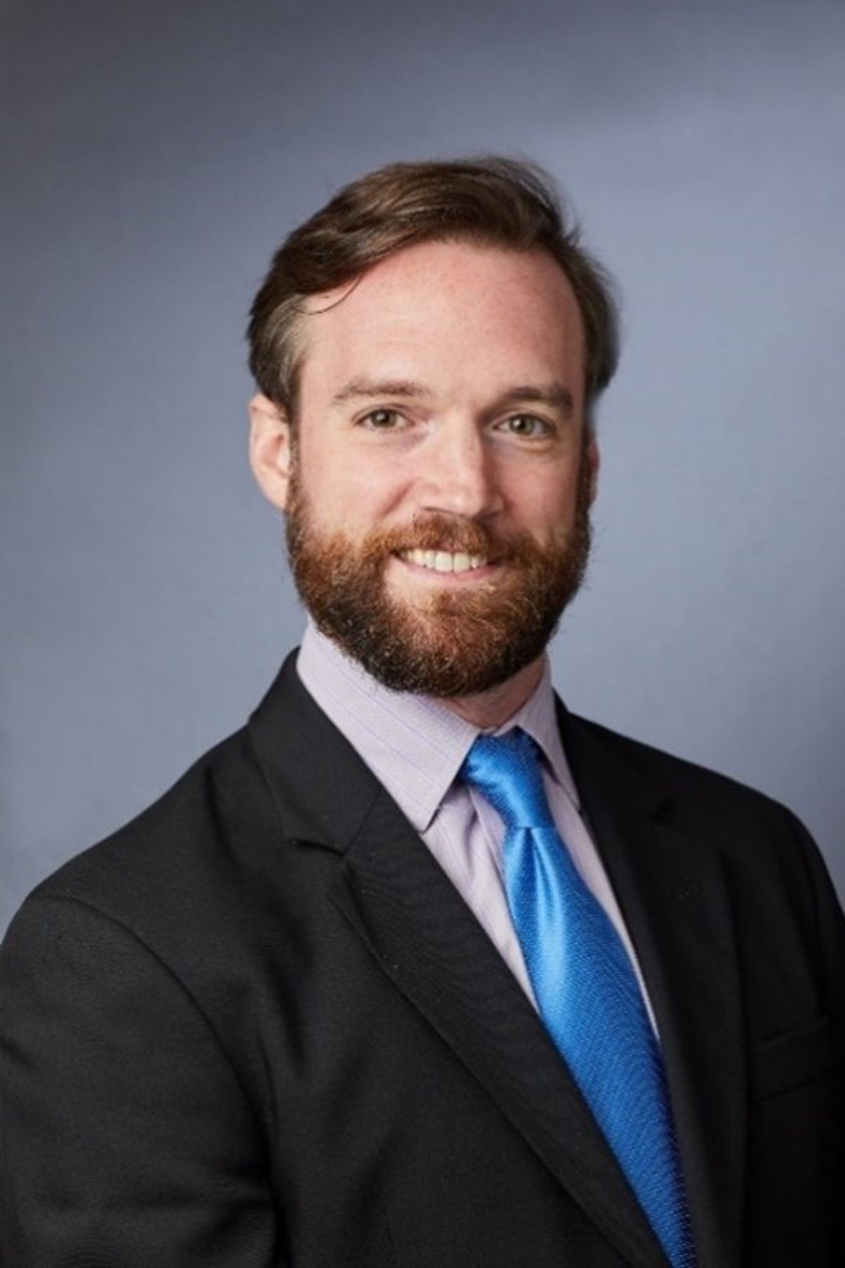 A headshot of Greg Tietjen, who is wearing a black suit and a blue tie.