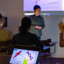 An image of a classroom where a scientist is giving a presentation using a slide deck projected on the wall.