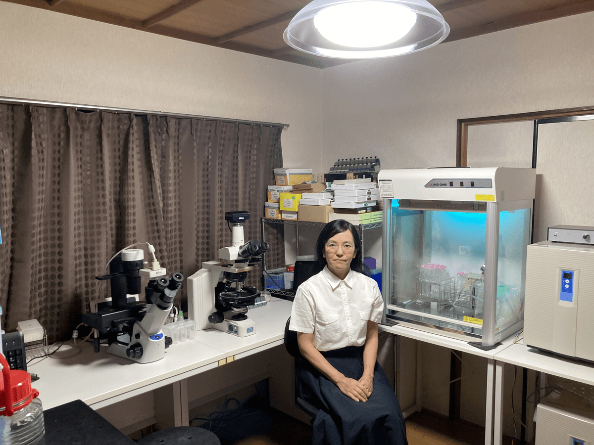 The researcher, Kyoko Hagino sits next to two microscopes, sterile hood, curtains, and she wears a blue skirt and white blouse.